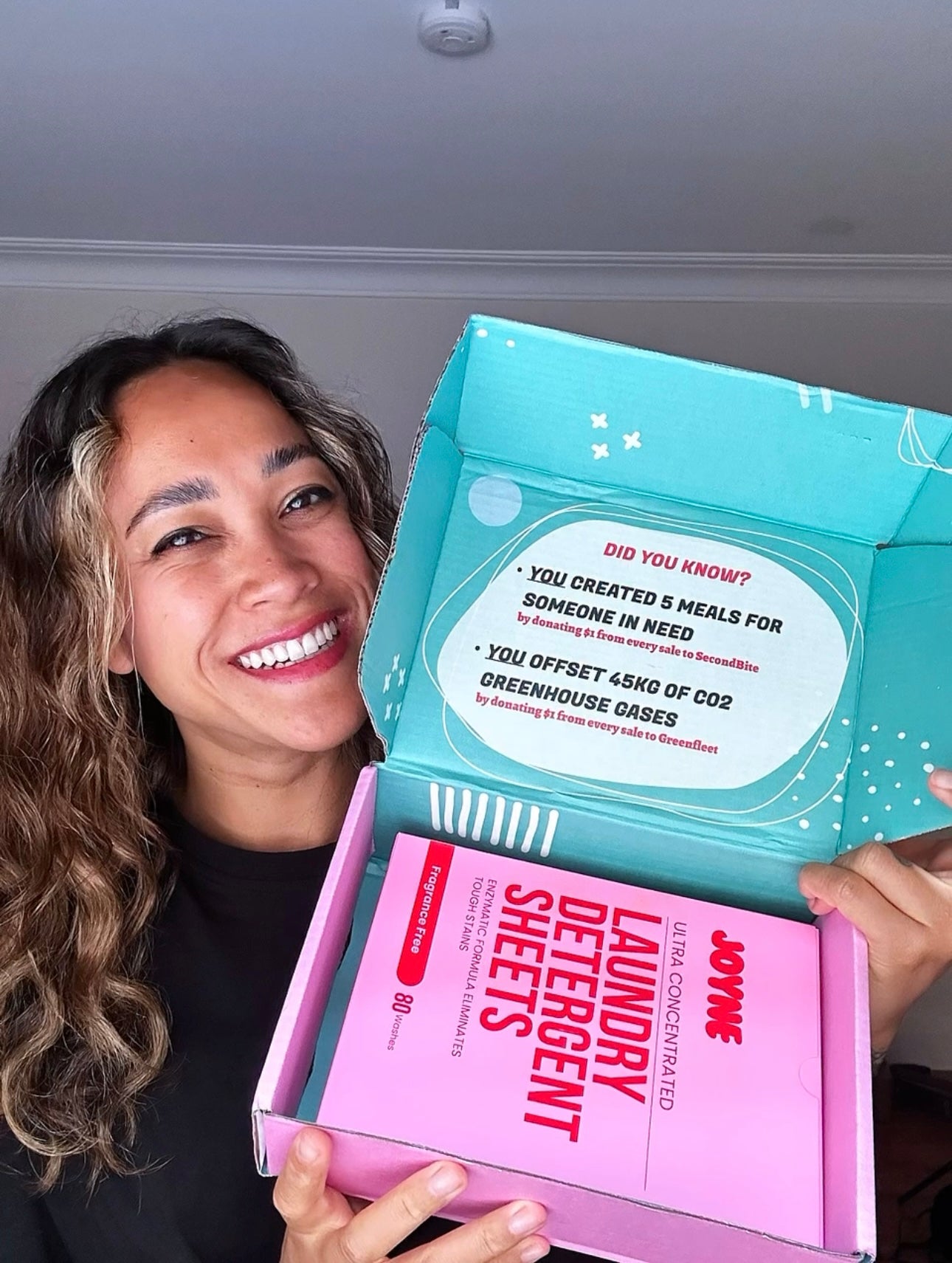 A smiling women holding an open box containing a pink box of laundry detergent sheets