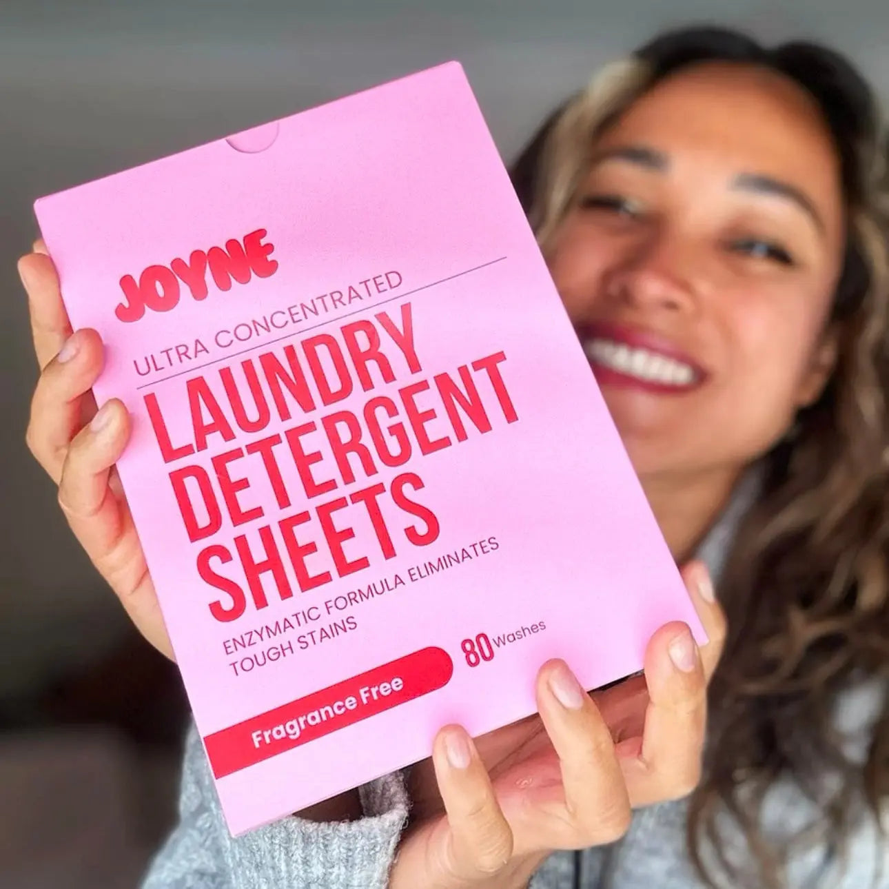 A female customer smiling and holding a pink box of JOYNE laundry detergent sheets