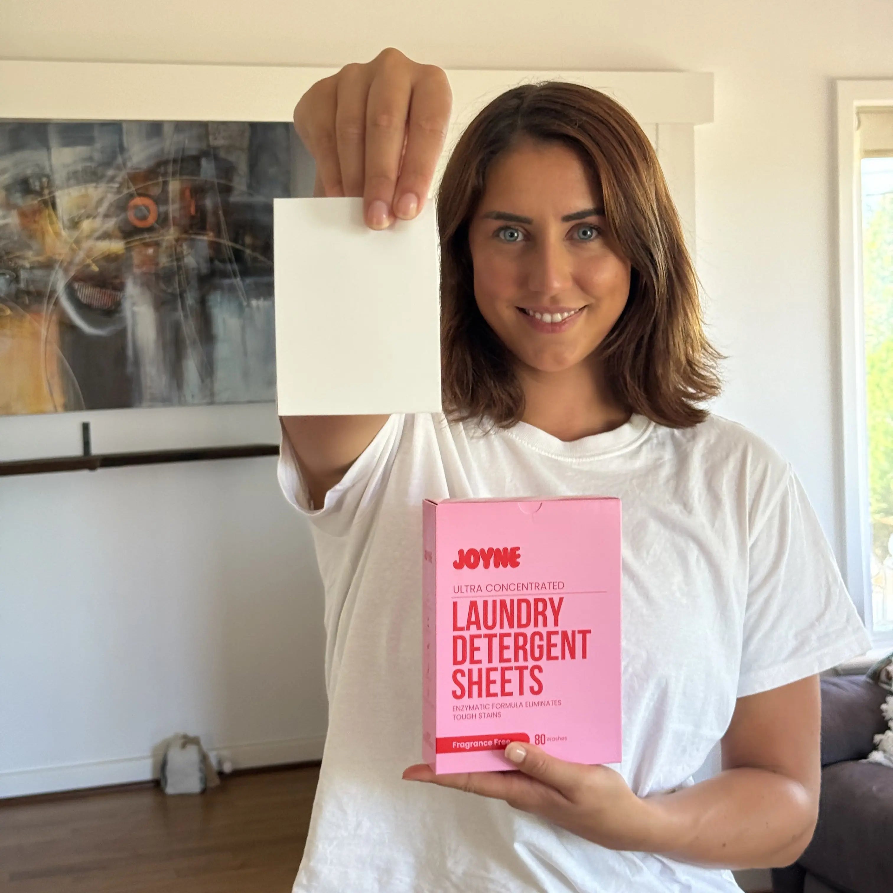 A female customer smiling and holding a JOYNE detergent sheet in one hand and a pink box of JOYNE laundry detergent sheets in the other