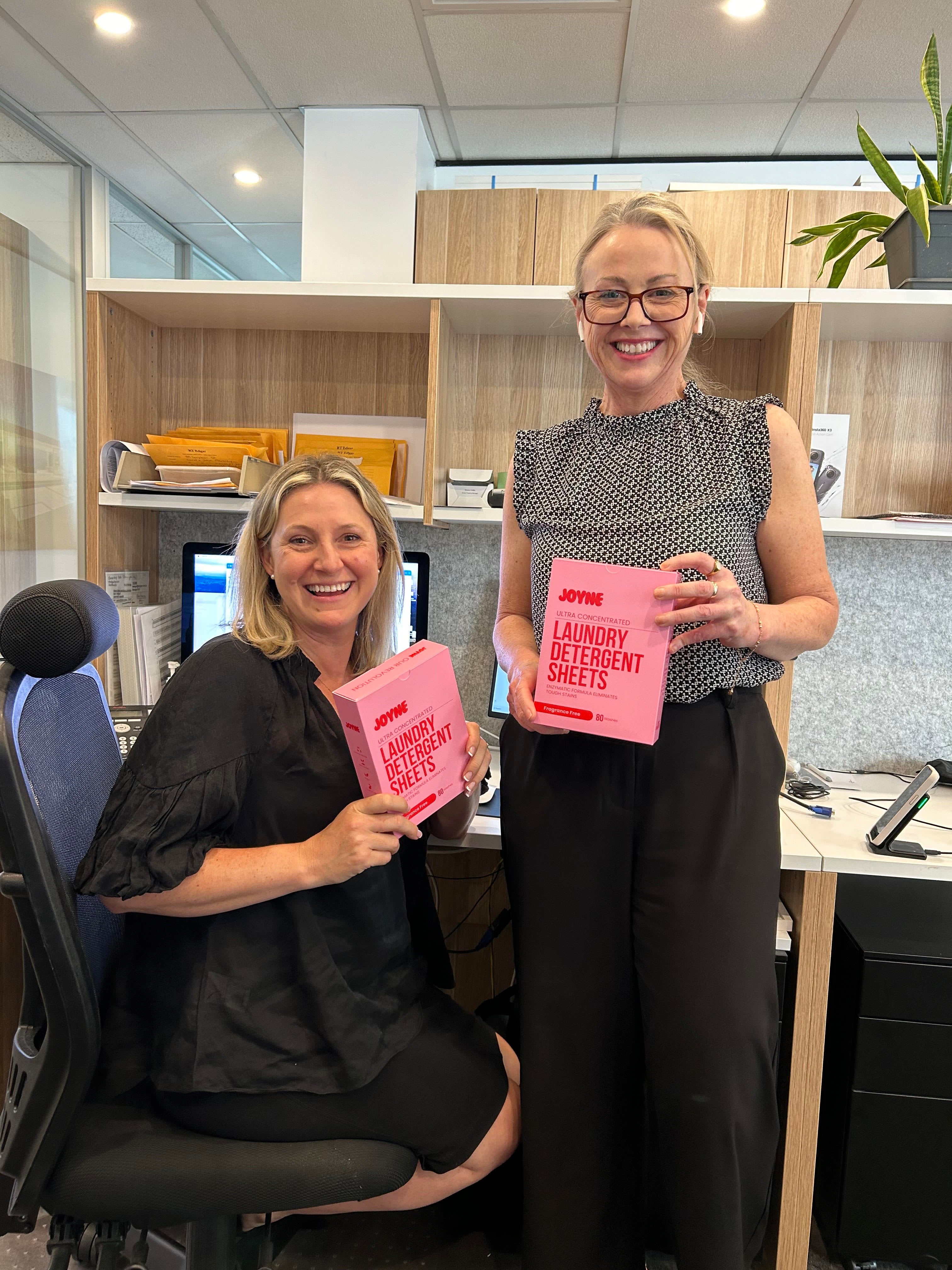 Two smiling women in a office holding a pink box of laundry detergent sheets